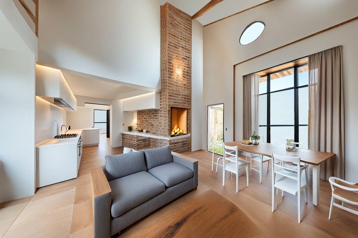 Spacious open-plan living and dining area featuring a striking double-height exposed brick fireplace, modern kitchen, and large windows providing abundant natural light.