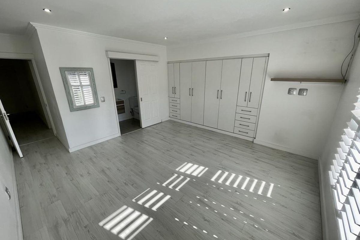 A bright, empty bedroom featuring modern laminate flooring, extensive built-in cabinetry, recessed lighting, and plantation shutters, with an adjoining en-suite bathroom.
