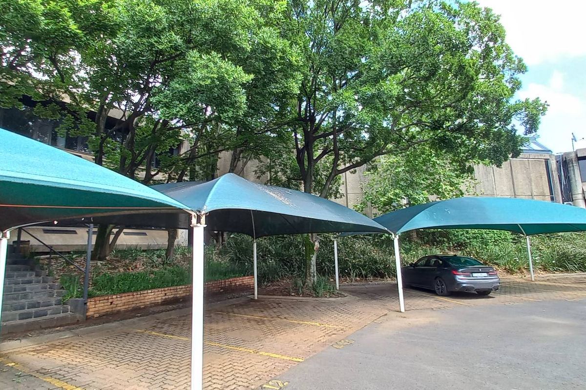 Outdoor parking area featuring multiple shade cloth structures, paved bays, and mature trees set against the office building facade.