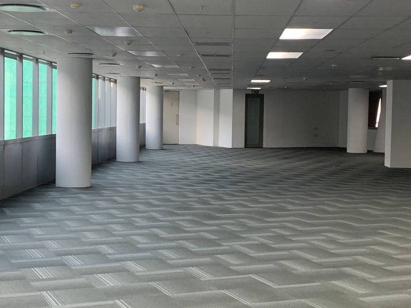 A spacious, open-plan office area featuring neutral carpet tiles, structural columns, and large windows providing ample natural light.
