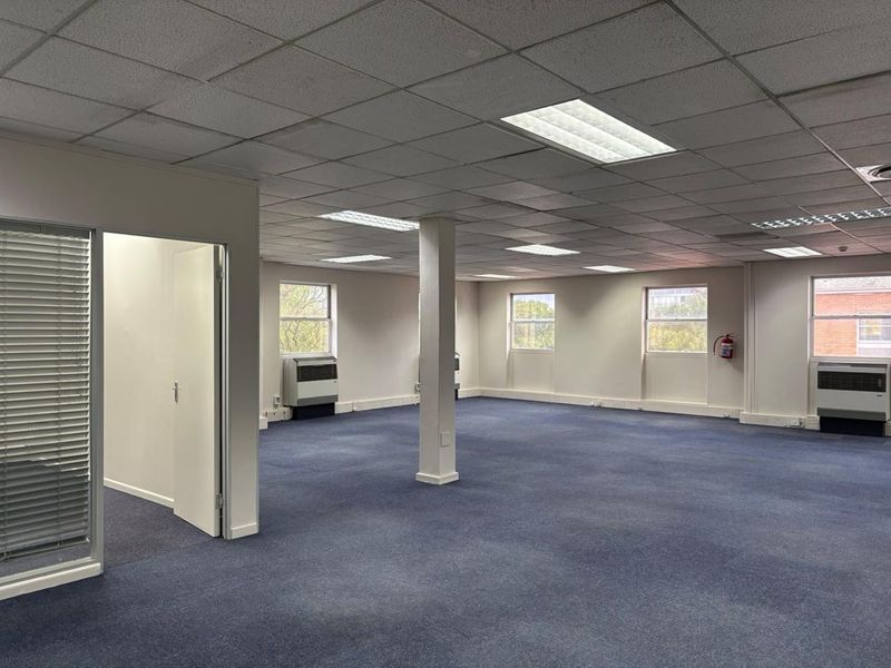 A spacious, open-plan office area featuring a suspended ceiling, recessed lighting, wall-mounted air conditioning units, and blue carpeting.