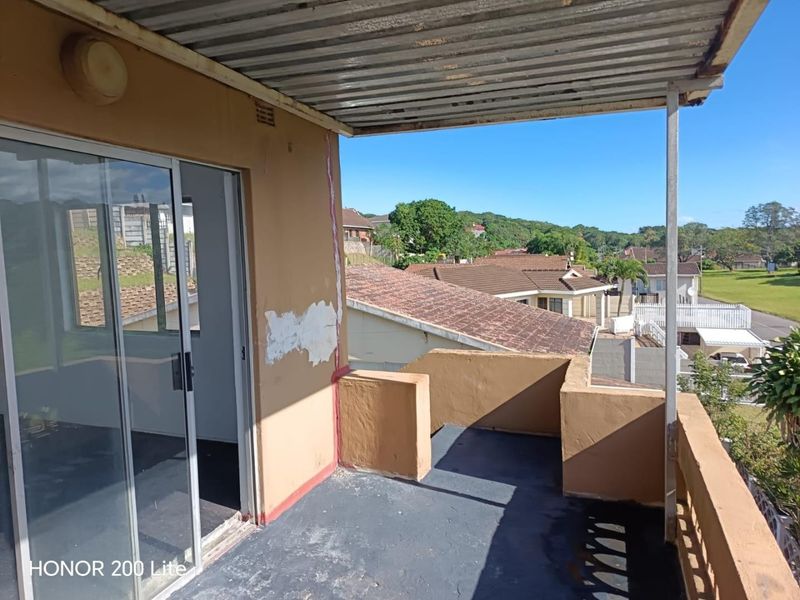 An elevated balcony featuring a corrugated metal roof, masonry balustrade, and sliding door access, offering views of the surrounding neighborhood.