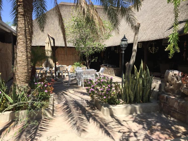 A covered outdoor patio area featuring a traditional thatched roof, stone paving, and established garden beds with a lamp post.