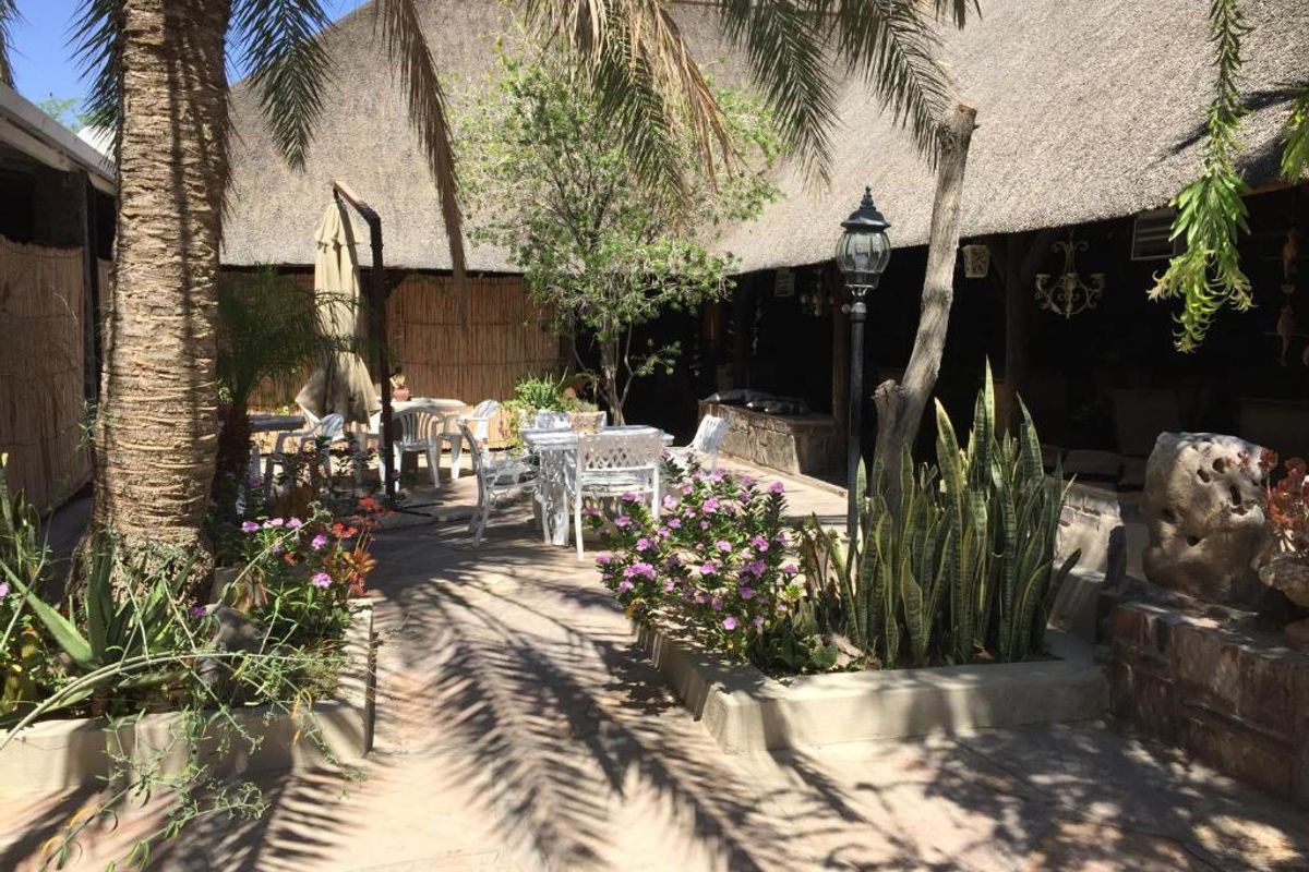 A covered outdoor patio area featuring a traditional thatched roof, stone paving, and established garden beds with a lamp post.