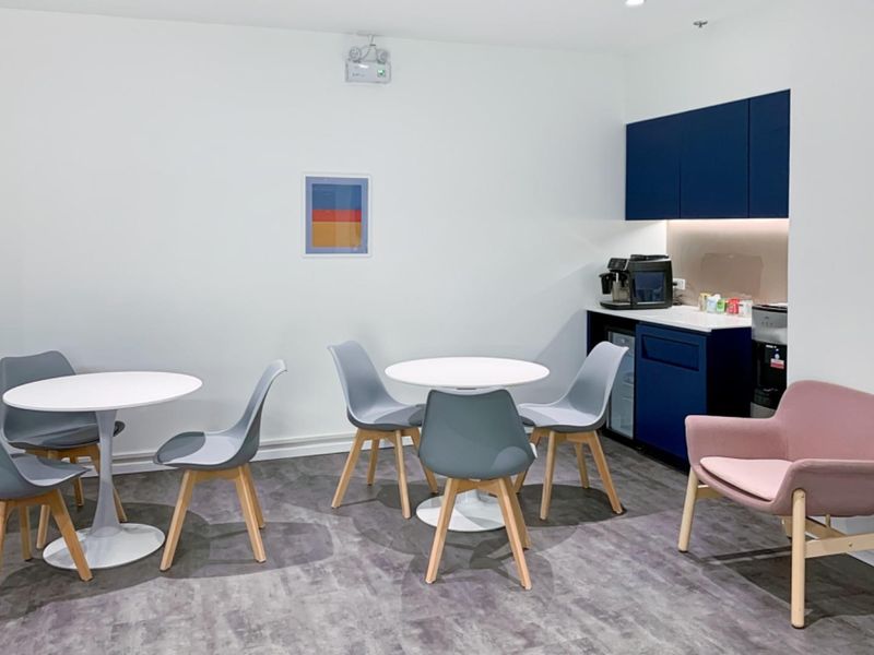 A modern office break area featuring a compact kitchenette with blue cabinetry, under-counter refrigeration, and comfortable seating arrangements for staff.