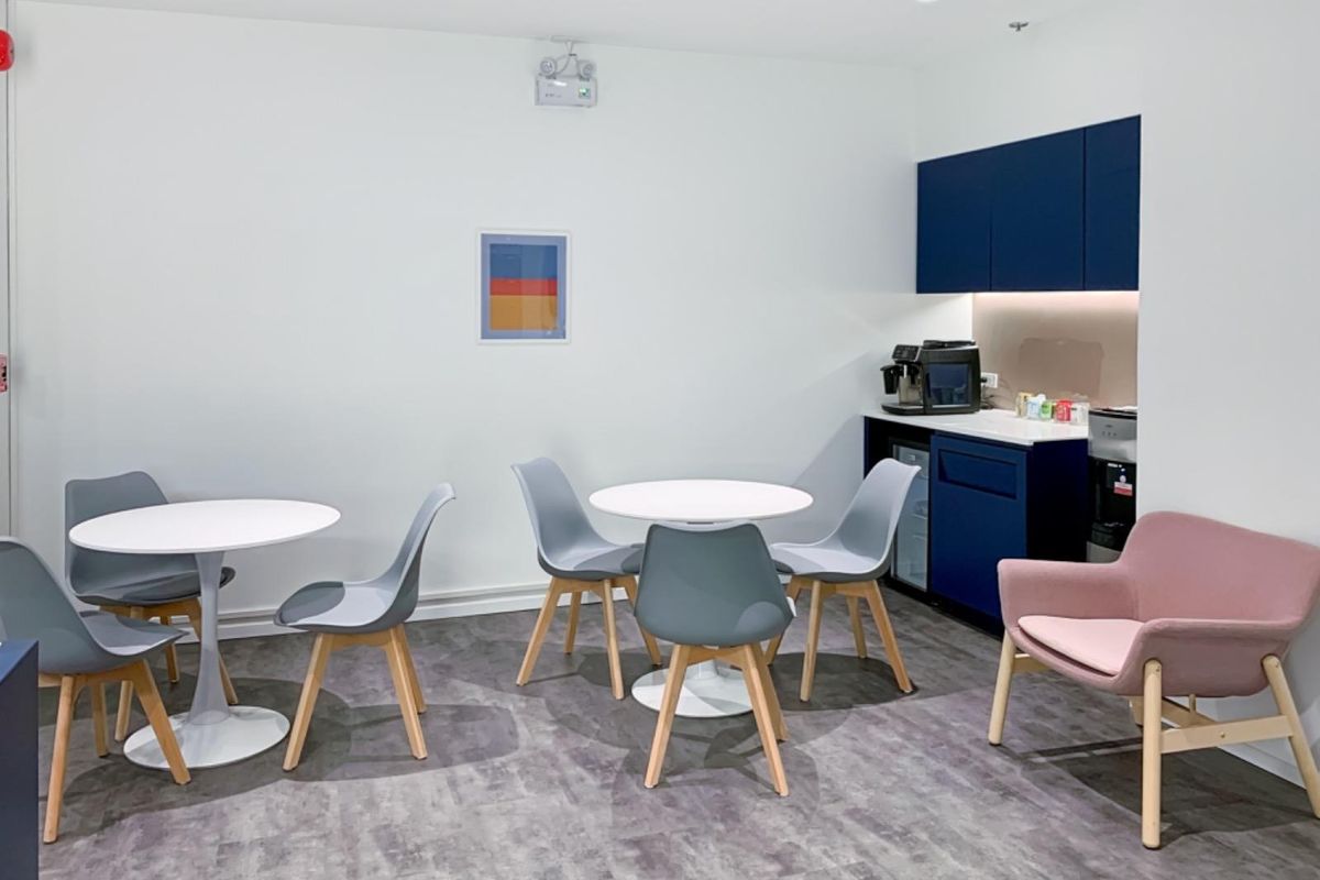 A modern office break area featuring a compact kitchenette with blue cabinetry, under-counter refrigeration, and comfortable seating arrangements for staff.