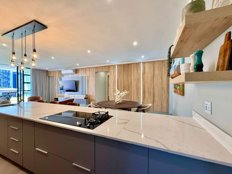 Modern open-plan living area featuring a sleek kitchen island with stone countertops, gas cooktop, and elegant timber wall paneling with integrated lighting.
