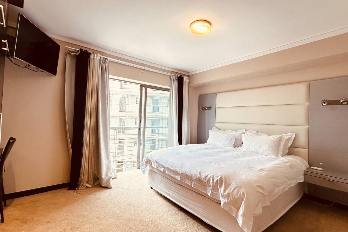 Spacious bedroom featuring a built-in headboard with integrated bedside tables, ample wardrobe storage, and a sliding glass door leading to a balcony.