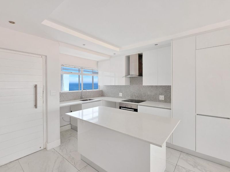 Modern, bright kitchen featuring white cabinetry, stone countertops, a central island, and integrated appliances with a scenic ocean view.