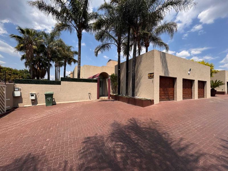 Front exterior view of a townhouse featuring a large paved driveway, triple garage with wooden doors, and established palm trees.