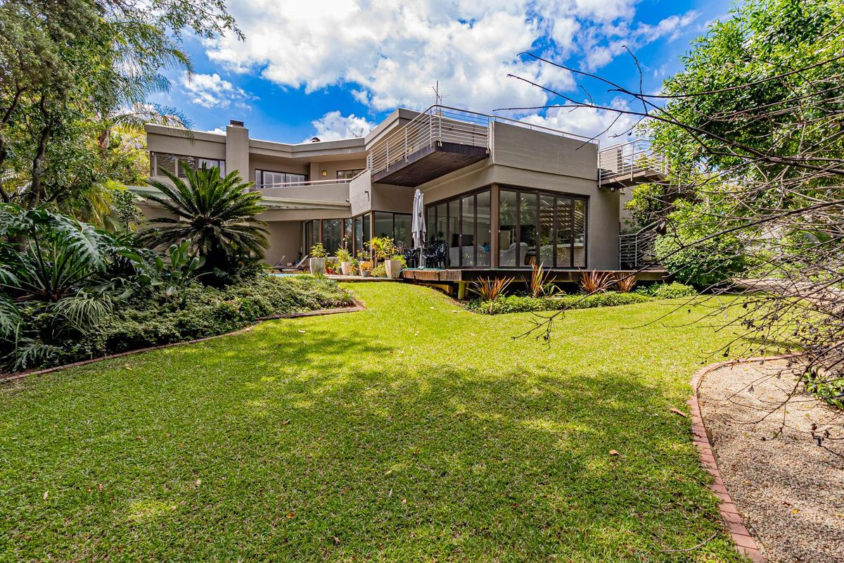 Rear view of a modern multi-level home featuring a spacious lawn, landscaped garden, wooden decking, and a balcony with metal railings.