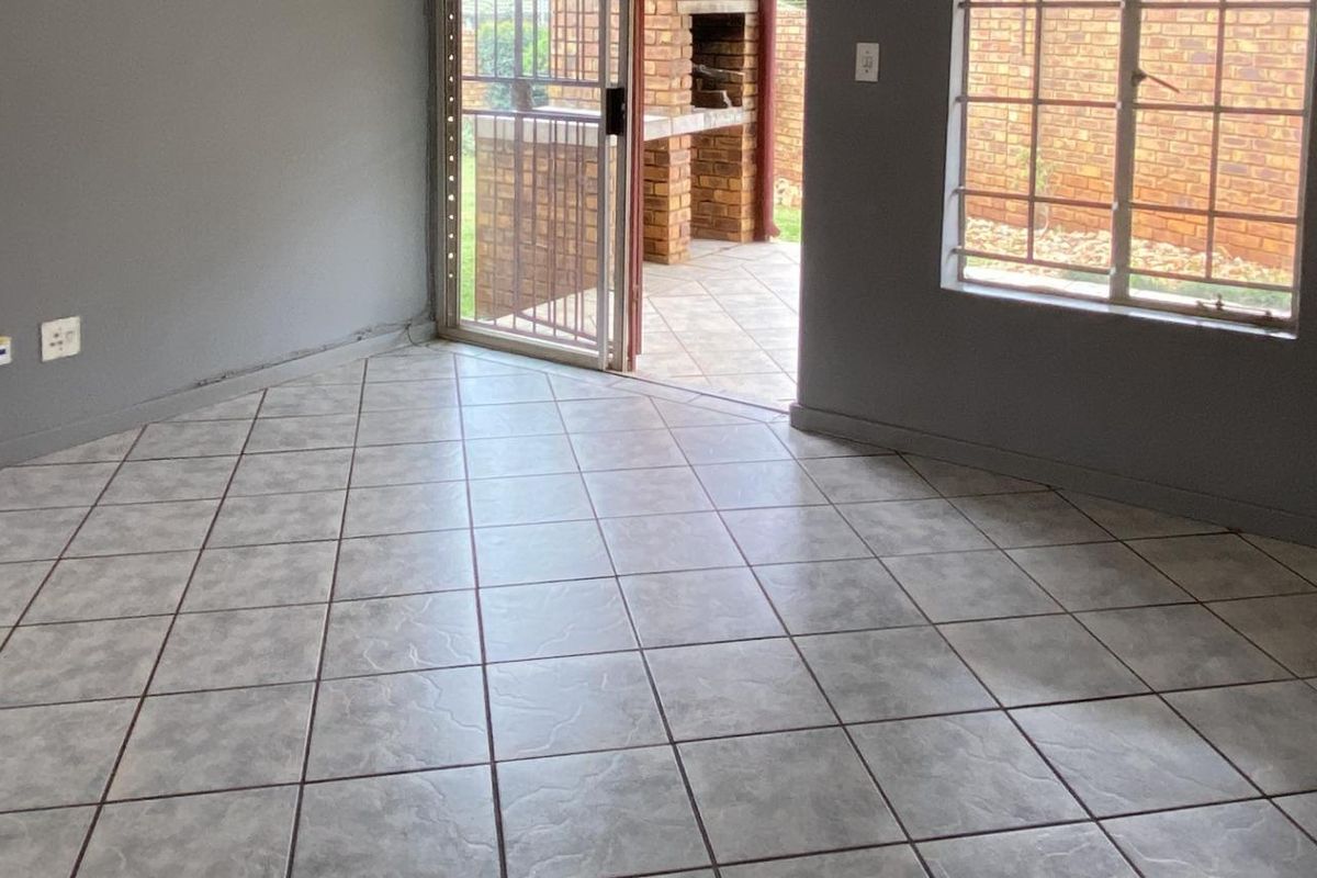 A bright living area featuring tiled flooring, large windows, and a sliding door providing direct access to an outdoor patio with a built-in braai.