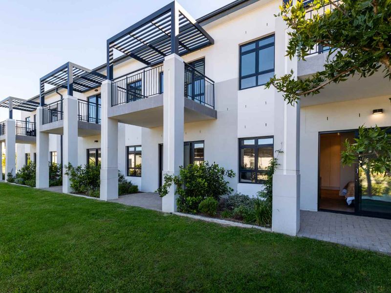 Rear elevation of a modern apartment complex featuring ground floor paved patios, upper level balconies with metal pergolas, and well-maintained lawn areas.