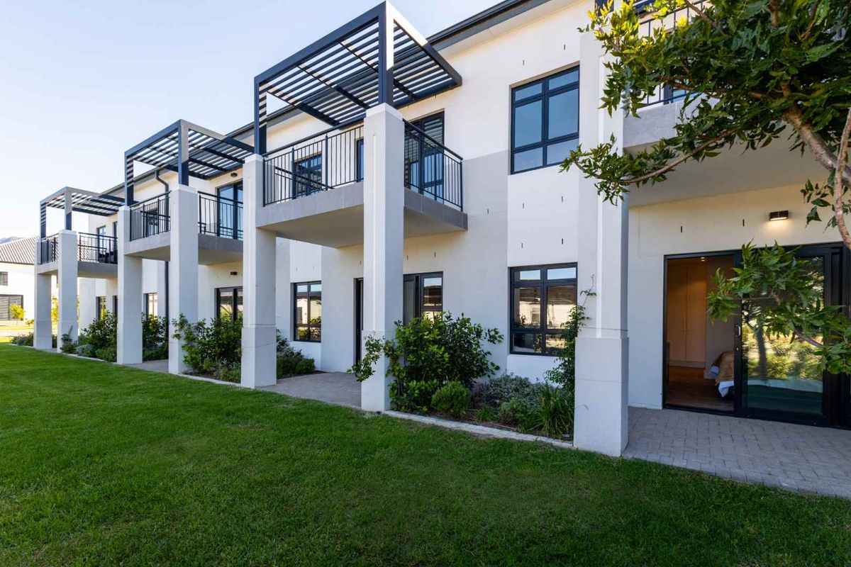Rear elevation of a modern apartment complex featuring ground floor paved patios, upper level balconies with metal pergolas, and well-maintained lawn areas.