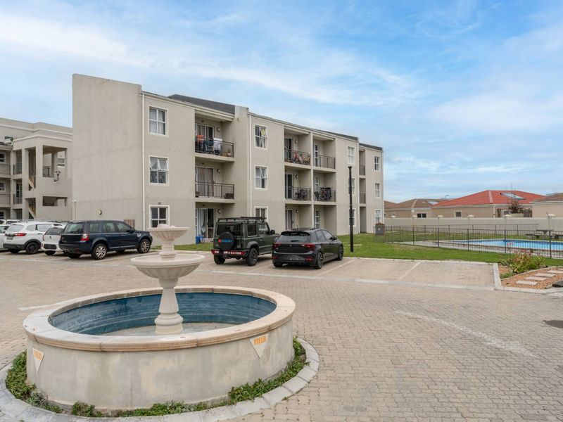 Exterior view of a multi-story apartment complex featuring a paved parking area, a central decorative fountain, and a communal swimming pool.