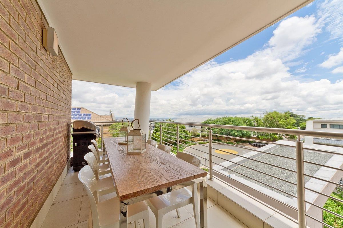 A spacious covered balcony featuring tiled flooring, a brick feature wall, and stainless steel railings, offering an elevated view of the surrounding neighborhood.