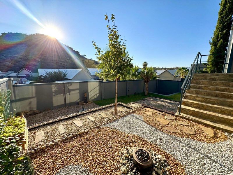 A well-maintained landscaped garden featuring gravel pathways, stone paving, a stone staircase with metal railings, and a solid boundary wall.