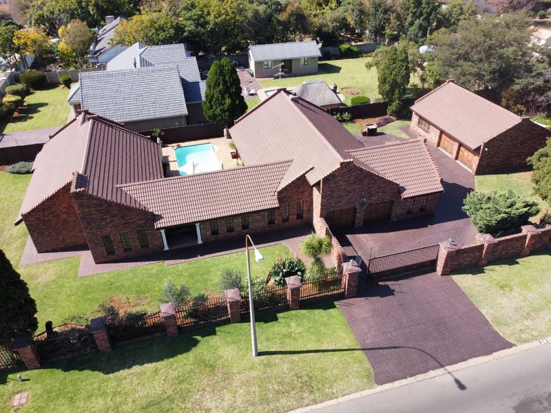 Aerial view of a spacious brick-faced residential property featuring a tiled roof, swimming pool, paved driveway, and detached garage set within a landscaped garden.