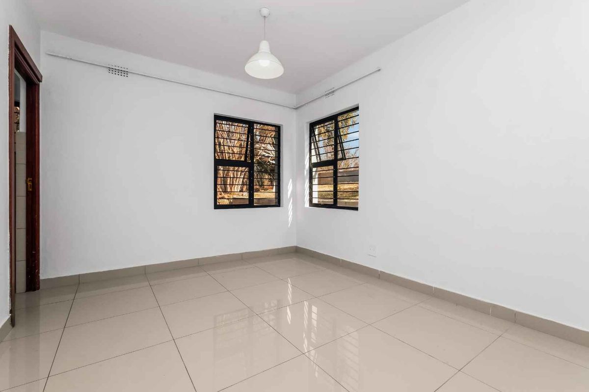 A bright, empty bedroom featuring neutral tiled flooring, white walls, and windows fitted with security bars.