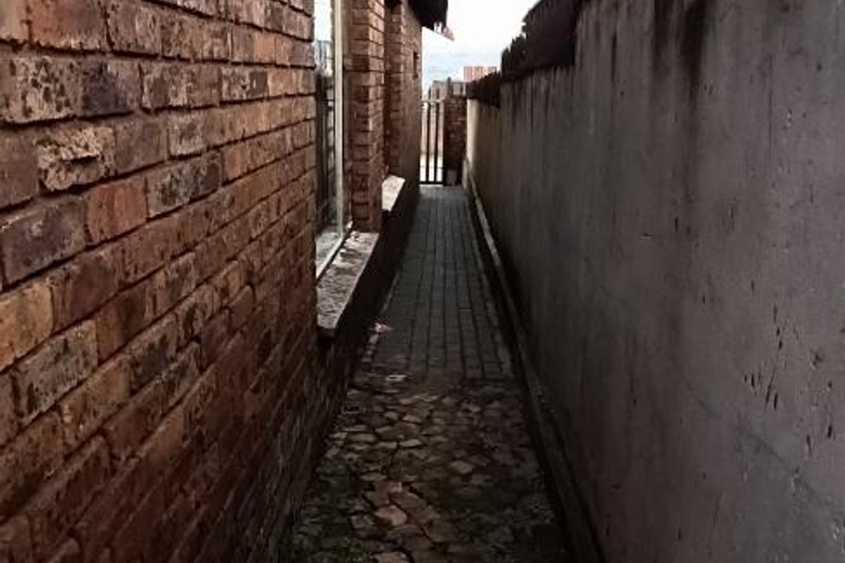 A narrow side passage featuring a brick exterior wall, paved walkway, and a high boundary wall with security spikes.