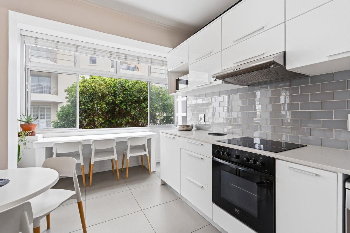 Modern kitchen featuring white cabinetry, stone countertops, subway tile backsplash, integrated oven, and hob, with a bright window-side breakfast bar area.