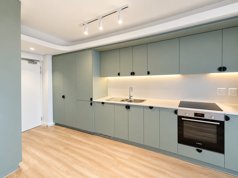 Modern kitchen featuring sage green cabinetry, integrated Bosch oven, electric hob, stainless steel sink, and sleek under-cabinet lighting with light wood-look flooring.