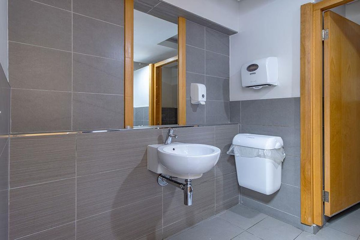 A clean, modern office restroom featuring grey wall tiles, a wall-mounted basin, and a wooden door frame.