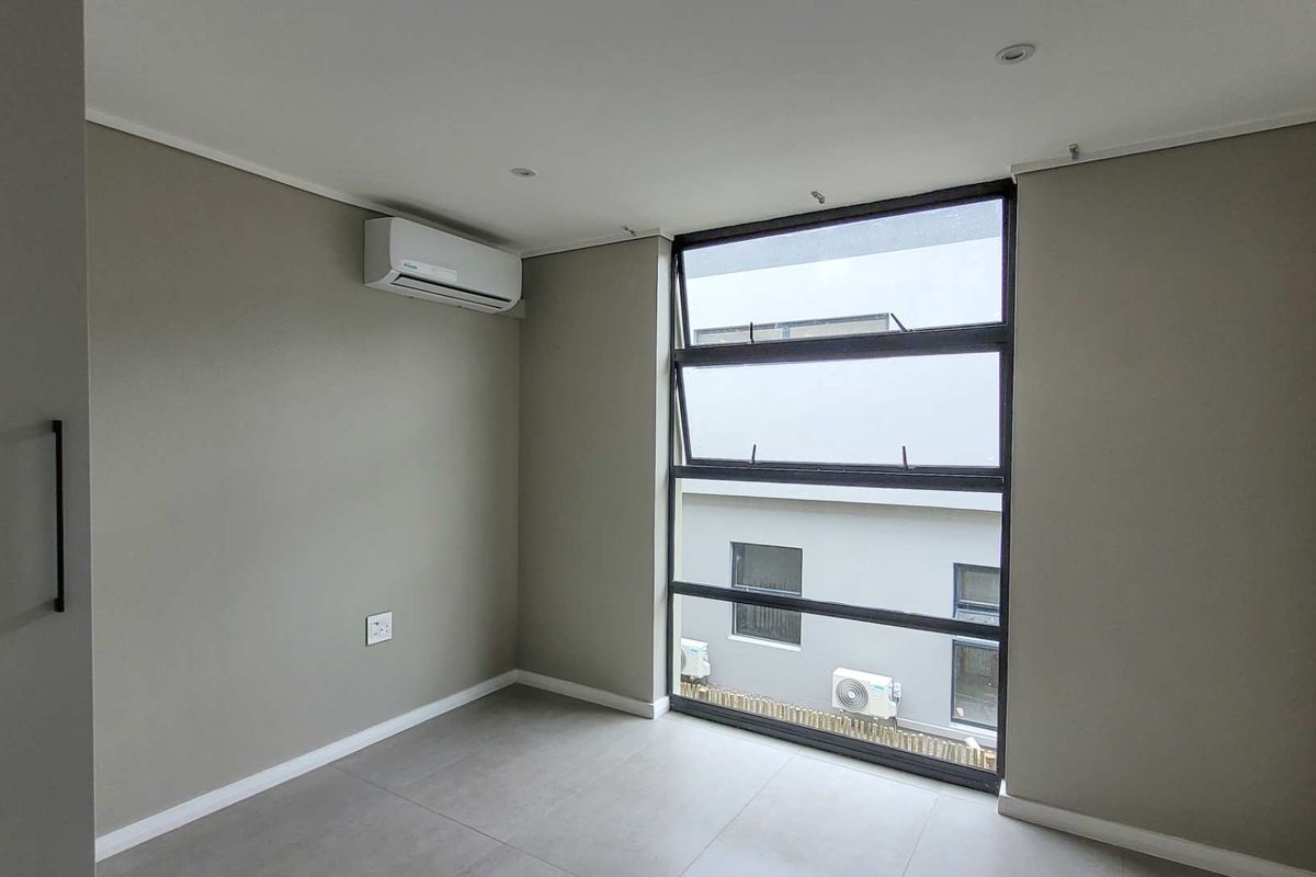 A bright, empty bedroom featuring neutral wall tones, large aluminium-framed windows, tiled flooring, and a wall-mounted air conditioning unit.