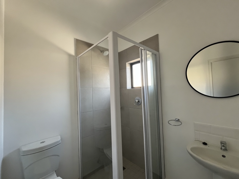 A clean and functional bathroom featuring a glass-enclosed shower, pedestal basin, and toilet with neutral wall tiling.