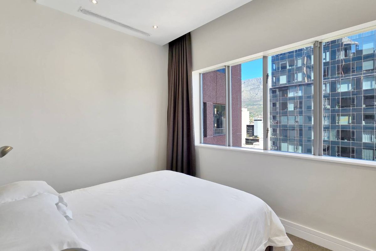 A bright bedroom featuring neutral walls, recessed lighting, ducted air conditioning, and large windows offering urban and mountain views.
