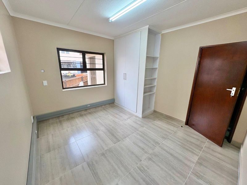 A clean, empty office space featuring neutral tiled flooring, built-in storage cabinetry, and natural light through a window with security bars.