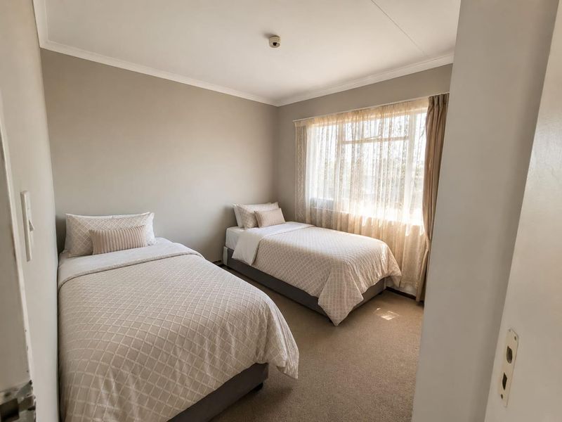 A well-lit bedroom featuring neutral wall tones, carpeted flooring, and a large window with sheer curtains, providing a comfortable and functional living space.