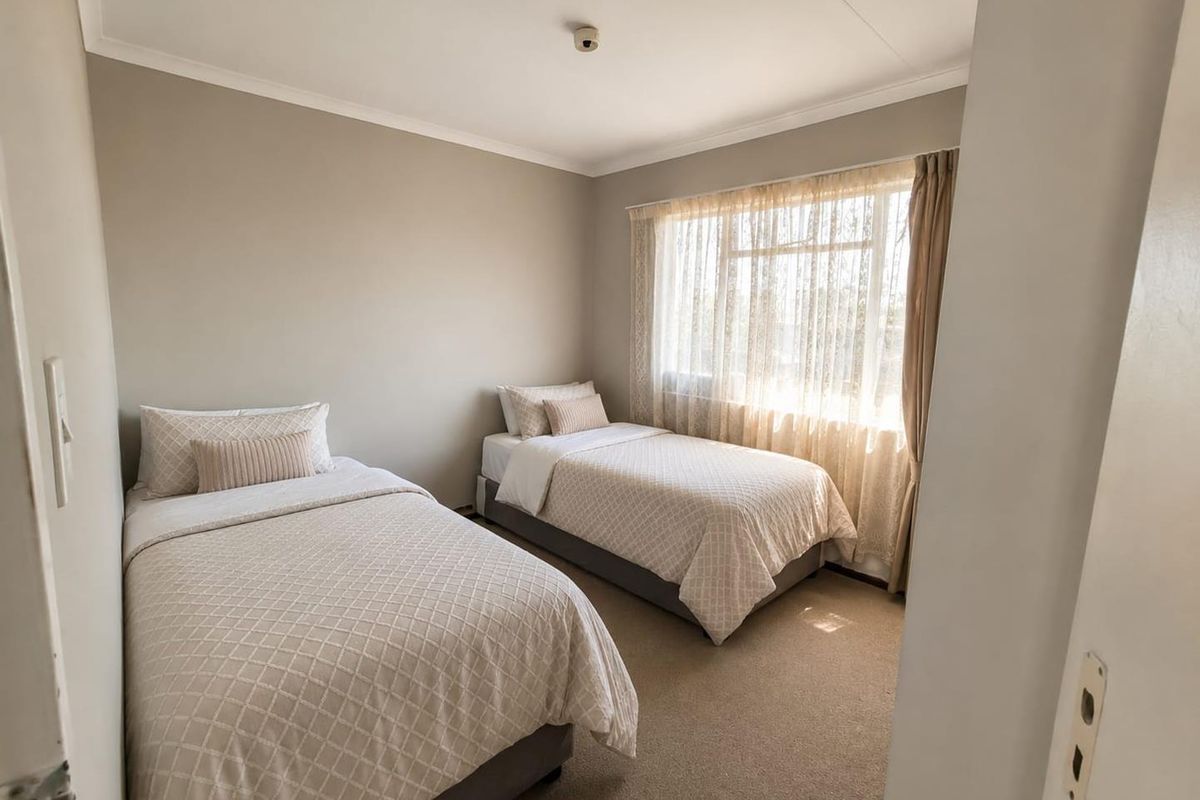 A well-lit bedroom featuring neutral wall tones, carpeted flooring, and a large window with sheer curtains, providing a comfortable and functional living space.