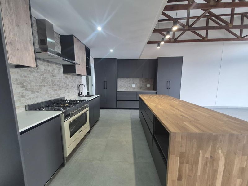 Modern kitchen featuring dark cabinetry, a large timber-topped island, gas stove, and striking exposed timber roof trusses in an open-plan layout.