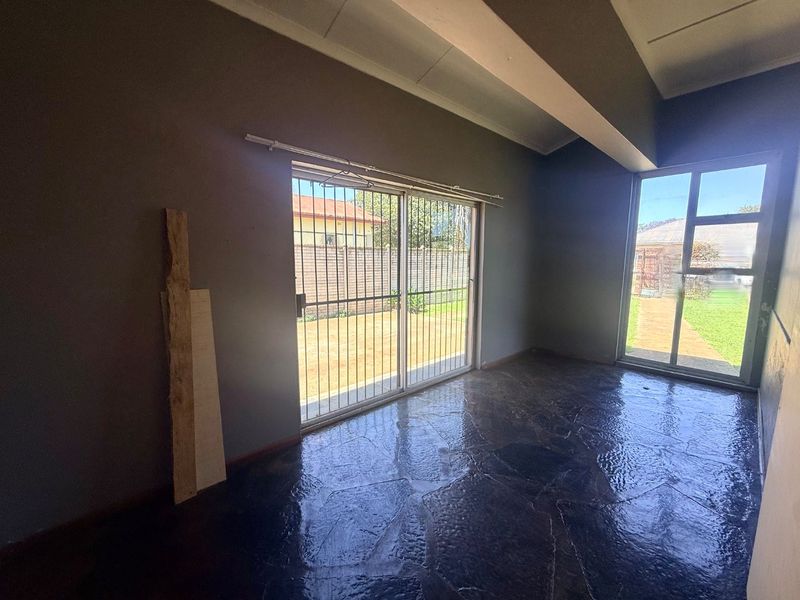 An empty living room featuring dark stone flooring, exposed ceiling beams, and large sliding glass doors with security gates leading to the garden.