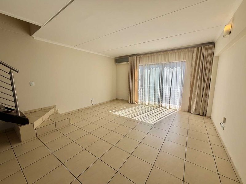 Spacious, light-filled living area featuring neutral tiled flooring, a staircase with modern metal railing, and direct access to a balcony through sliding glass doors.