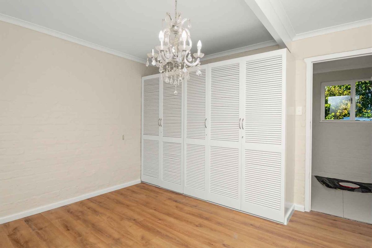 A bright, empty bedroom featuring white louvered built-in wardrobes, wood-look laminate flooring, and a decorative crystal chandelier.