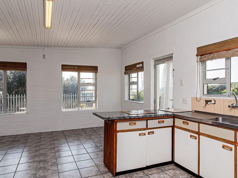 An empty, dated kitchen featuring laminate cabinetry, granite-look countertops, ceramic floor tiles, and ample natural light through multiple aluminium-framed windows.