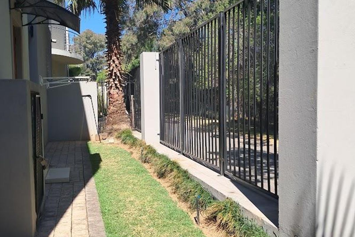 A side garden area featuring a paved walkway, manicured lawn, and secure metal perimeter fencing with concrete boundary walls.