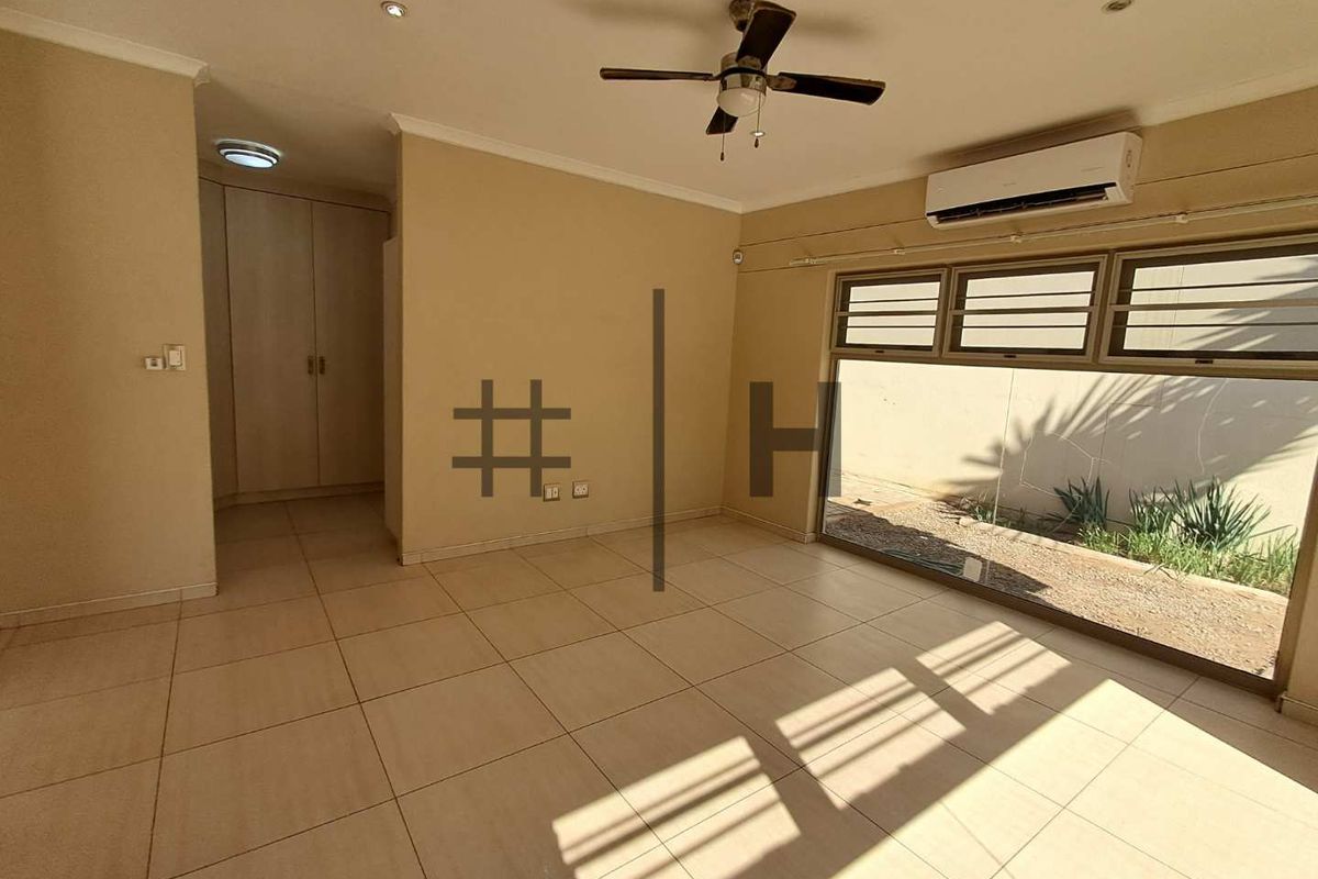 Spacious living area featuring tiled flooring, built-in cupboards, a ceiling fan, and a wall-mounted air conditioning unit with large windows overlooking the exterior.