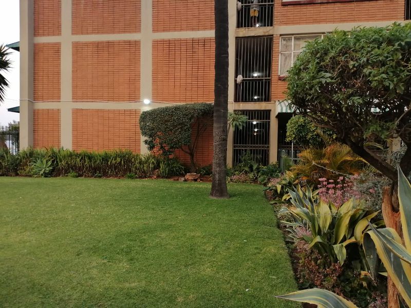 A view of the apartment building exterior featuring a well-maintained lawn, landscaped garden beds, and brick facade with security-barred windows.