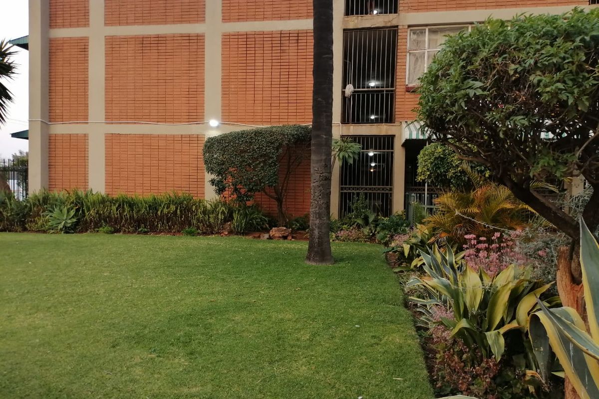 A view of the apartment building exterior featuring a well-maintained lawn, landscaped garden beds, and brick facade with security-barred windows.