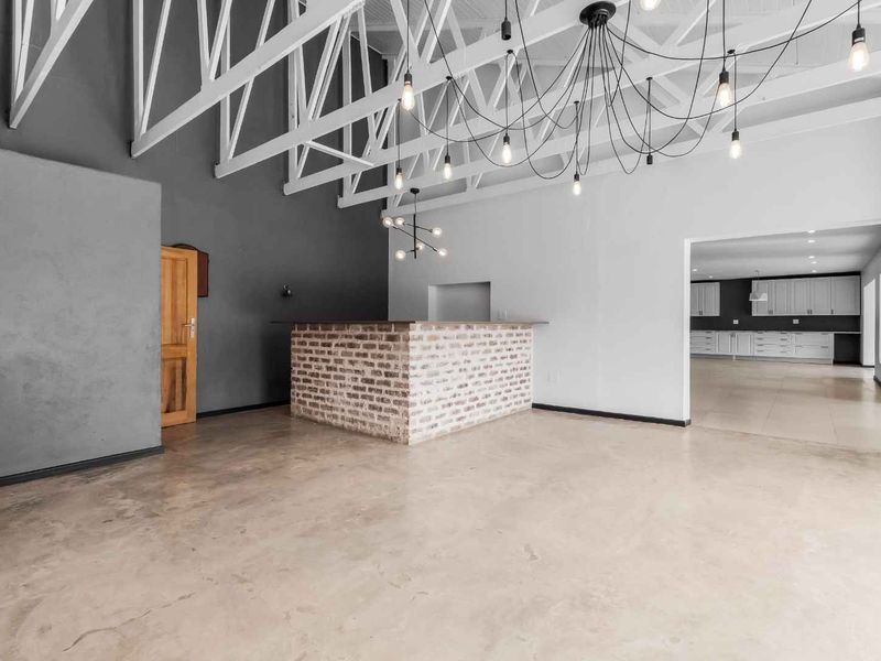 Spacious open-plan living area featuring exposed white roof trusses, polished concrete floors, a central brick bar, and an adjoining kitchen space.
