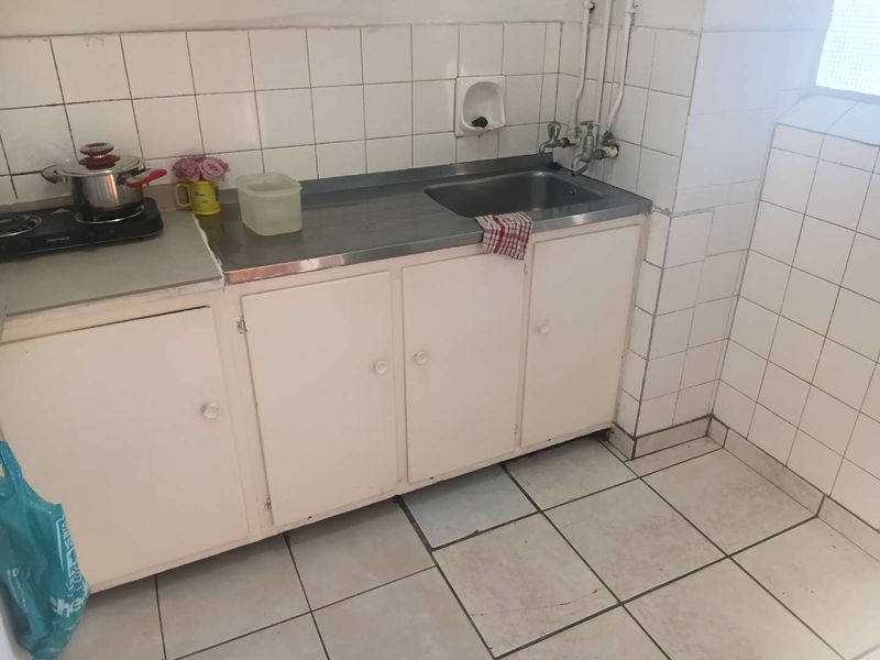 A dated kitchen area featuring a stainless steel sink, tiled backsplash, and floor tiles, with visible wear on the cabinetry and exposed plumbing.