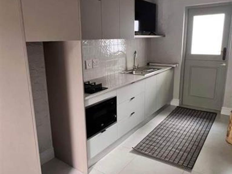 A modern, clean-lined kitchen featuring neutral cabinetry, built-in appliances, tiled backsplash, and exposed white roof trusses, leading to an exterior door.