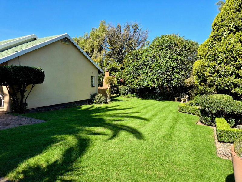 A well-maintained rear garden featuring a lush lawn, mature trees, manicured hedging, and a brick chimney structure attached to the side of the house.