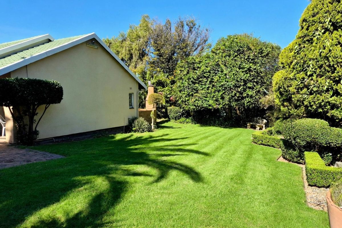 A well-maintained rear garden featuring a lush lawn, mature trees, manicured hedging, and a brick chimney structure attached to the side of the house.