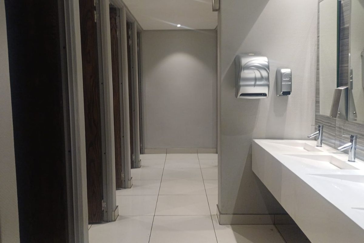 A clean, modern office bathroom facility featuring multiple toilet cubicles, a long integrated vanity basin, and wall-mounted dispensers.