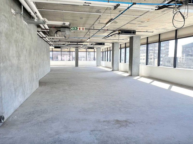 An expansive, open-plan office space featuring an industrial aesthetic with exposed concrete ceilings, ducting, and large windows providing ample natural light.