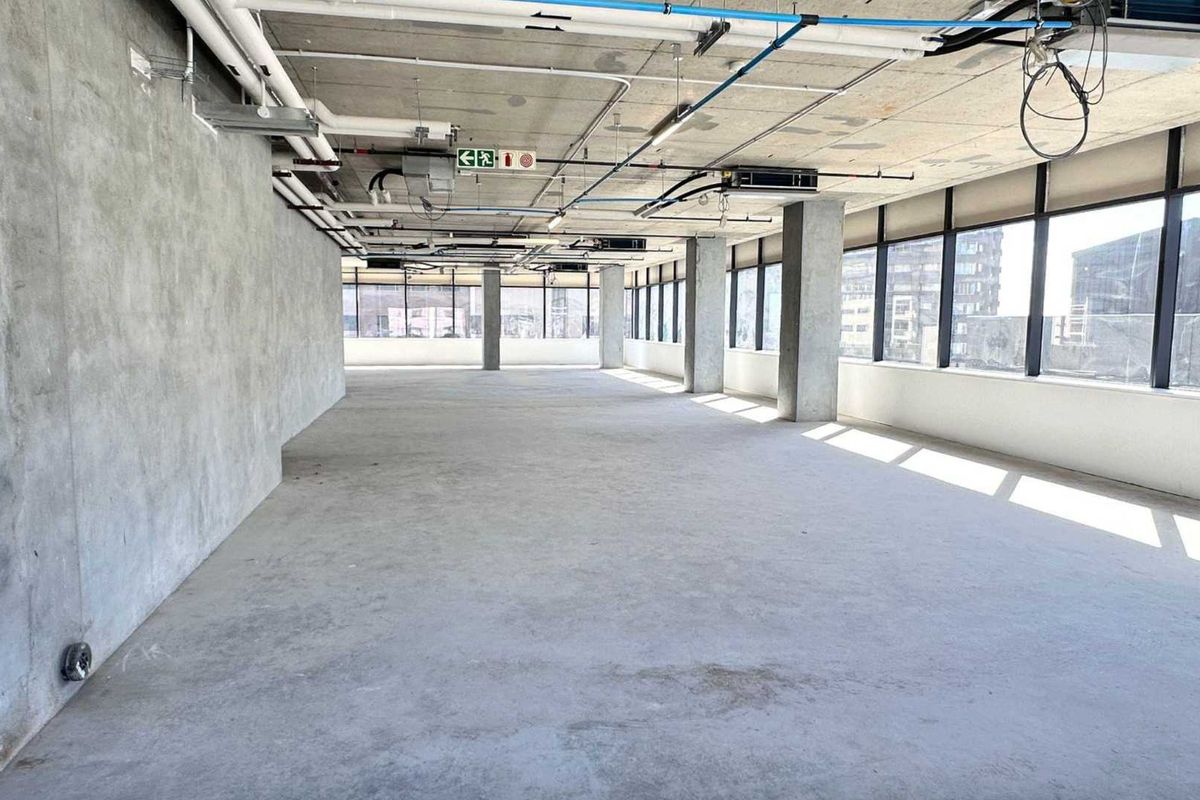 An expansive, open-plan office space featuring an industrial aesthetic with exposed concrete ceilings, ducting, and large windows providing ample natural light.
